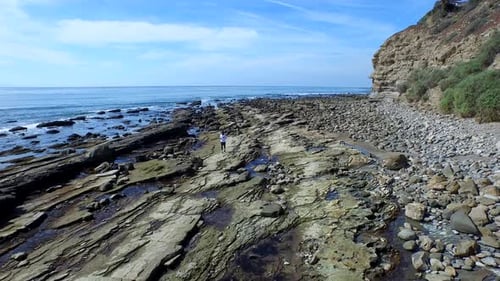 Tracking shot of a young man running on a rocky ocean beach shoreline.