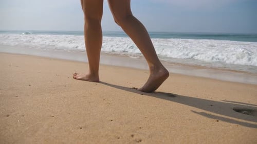 Female Feet Walking on Golden Sand at the Beach with Ocean Waves at Background
