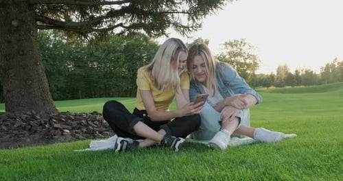 Two Girlfriends Sitting on Green Grass of Lawn Smiling Talking Looking Together at Smartphone Screen