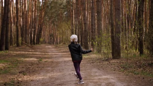 The Little Girl Runs Along the Path Road in the Pine Forest. Dressed in the Leather Jacket.