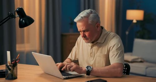 Elderly Man with Gray Hair Works on Laptop at Desk and Lamp in Evening