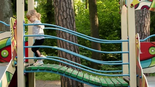 Cheerful Blonde Kid Girl with Ponytail in Orange Round Glasses Plays on Playground in Park at Sunset
