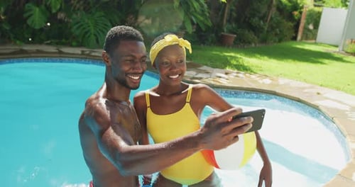 Happy african american couple standing in swimming pool taking selfie and smiling in sunny garden