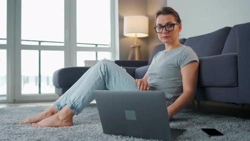 Woman With Laptop Sits on Carpet Leaning on Couch