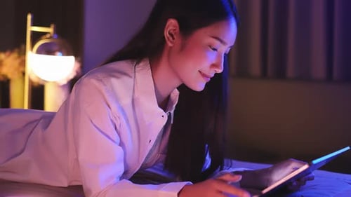 Young Asian beautiful woman enjoy using smartphone- tablet in bedroom at night time.