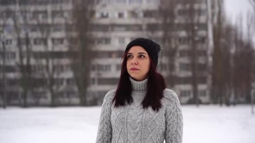 Thoughtful Young Woman in Gray Knitted Sweater and Hat Standing on Street in Winter Season