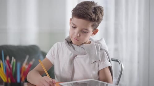 Portrait of Concentrated Caucasian Boy Sitting at the Table and Drawing or Writing in Notebook