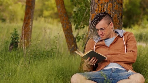 Ein Mann in Freizeitkleidung und Brille liest Literatur unter einem Baum
