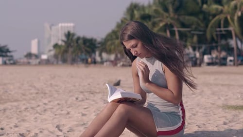 Young Woman with Dark Hair Sits on Sandy Beach and Reads