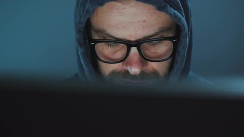 Hacker masculino en el capó y gafas trabajando en una computadora en una sala de oficina oscura