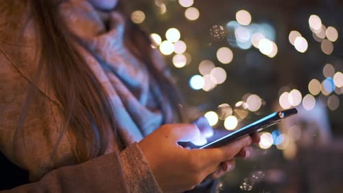 Close-up of Woman Uses a Smartphone in Winter on the Background of a Christmas Tree Decorated with