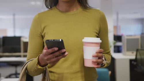 Biracial businesswoman smiling and walking with smartphone in office