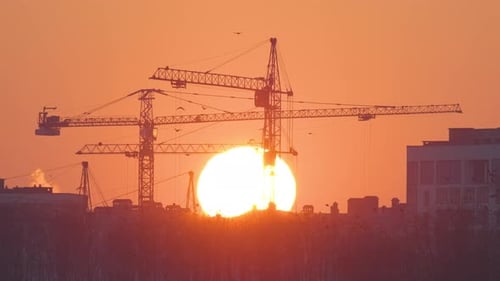 Dark Silhouette of Tower Cranes with Big Setting Sun at High Residential Apartment Buildings