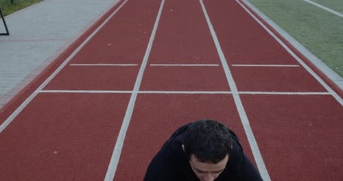 Disabled Man with Prosthetic Running Blades Stretching Back While Sitting at Track on Sports Field