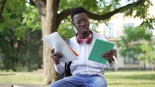 Zoom in to Stressed Overloaded Young Student Sitting on Bench with Tablet and Paperwork Holding Head