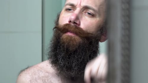 Bold Man Is Combing His Beard and Mustache. Attractive Man in Bathroom Looks in Mirror and Combes