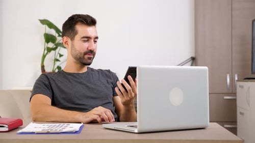 Young Man Using Phone at Desk with Laptop
