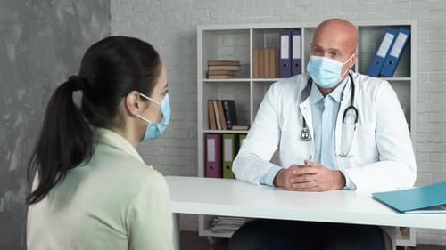 Mature Doctor Welcoming Female Patient, Covering Face with Medical Masks, Consulting in a Clinic