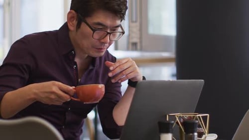 Asian man with laptop drinking coffee while sitting at a cafe