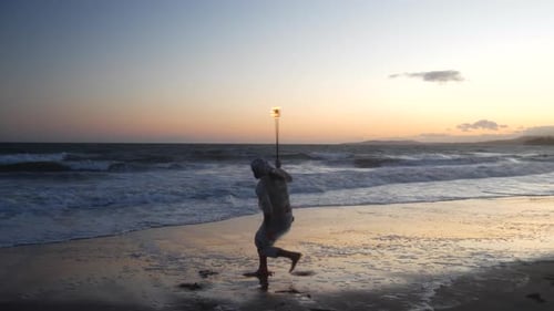Ein Mann Feuertänzer in Silhouette dreht seinen flammenden Stab am Strand bei Sonnenuntergang mit Flammen und Oc