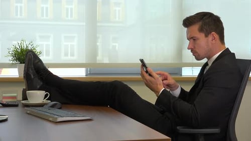 An office worker in a suit sits in front of a computer with legs on desk and works on a smartphone