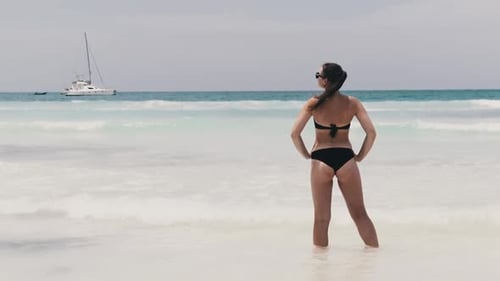 Young Woman in Bikini Standing on a Beach and Looks at Turquoise Ocean