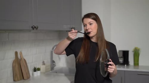 Woman Tastes Food with Spoon in Kitchen