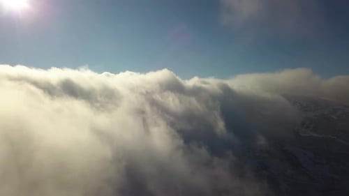 Aerial view from above of white puffy clouds covering snowy mountain tops in bright sunny day.