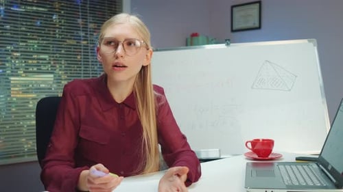 Woman Lecturing at Desk with Equations on Whiteboard