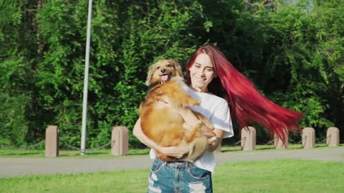 Happy Beautiful Woman with Red Hair Spin with Dog in Hands on Green Lawn in Forest. Love for pets