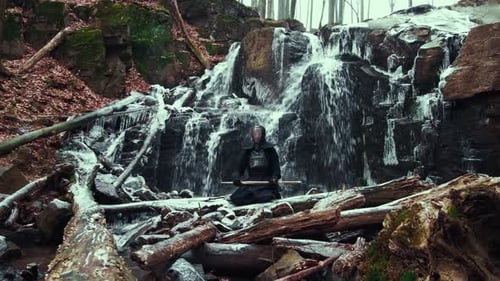 Man in Japanese Uniform Sits By Icy Waterfall
