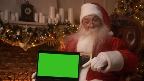 Santa Claus Holds a Laptop with Green Screen and Points at It Chroma Key While Sitting in His Rocker