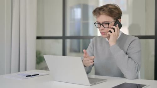 Young Man Talking on Phone While Using Laptop in Modern Office