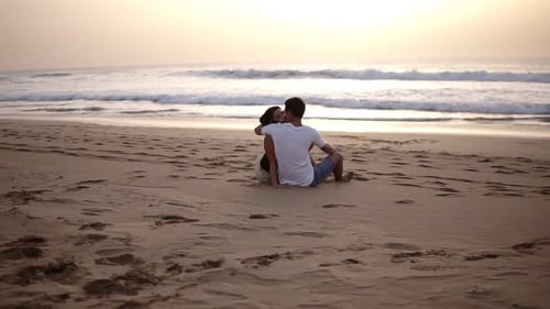 A Couple Sit in the Sand at the Beach Looking Out to the Ocean and Kiss in the Sunset