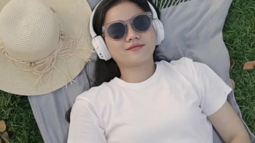 Lächeln einer asiatischen Frau mit Sonnenbrille und Kopfhörern, während sie auf dem Bodengras liegt.