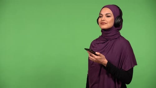A Muslim Woman Listens to Music with Headphones on and a Smartphone and Smiles Green Screen