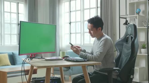 Asian Man With Green Screen Desktop Computer Using Mobile Phone While Working At Home