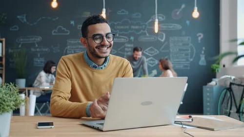 Cheerful Arab Man Talking Online Using Laptop Computer Looking at Camera in Open Space Office