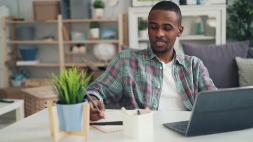 Afroamerikaner arbeitet zu Hause mit Laptop, schreibt dann Notizen auf Papier und trinkt
