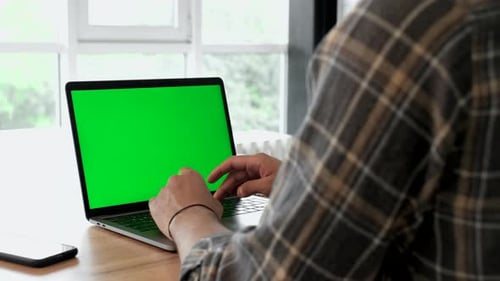 Close-up of a Man Uses Laptop with Green Mock-up Screen While Sitting at the Desk in His Cozy