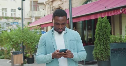 Cara de pele negra em roupas casuais que anda perto de um café de rua e usa o celular