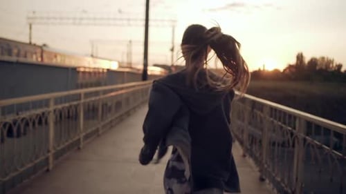 View From Back Young Sporty Girl with Fluttering Hair Running Along City Bridge at Sunset with