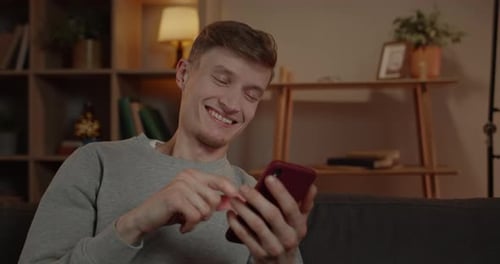 Close Up View of Millennial Man Scrolling Smartphone Screen and Smiling in Living Room