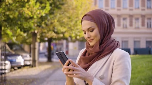 A Young Beautiful Muslim Woman Takes Selfies with a Smartphone in a Street in an Urban Area