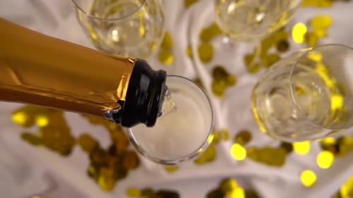 Pouring from a bottle of sparkling wine into glasses on a background of confetti. Slow motion.
