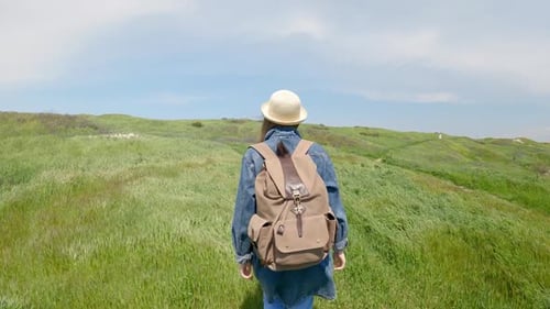 A Woman in a Hat with a Large Backpack Walks Along a Green Meadow with Green Grass