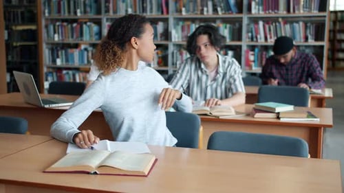 African American Girl Studying in Library Reading Taking Book From Male Student