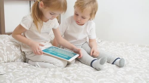 Girl and Boy Play on a Tablet on Bed