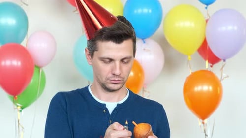 Positive Man Burns Candle on Small Cupcake Against Balloons