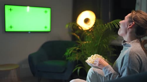 Rear View of a Woman Sitting on a Sofa in the Living Room in the Evening and Watching a Green TV